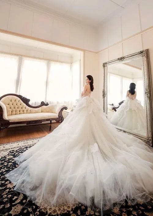 A bride in a voluminous white wedding gown stands in front of a large ornate mirror in an elegant, well-lit room. There is a cream-colored vintage sofa by the window, and a black and gold patterned rug covers the wooden floor.