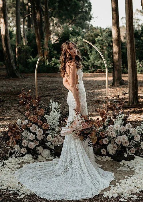 A woman wearing a lace wedding dress stands outdoors among trees. She holds a large bouquet of flowers and stands on a circular arrangement of white rose petals. An elegant floral arch with gold accents frames her from behind.