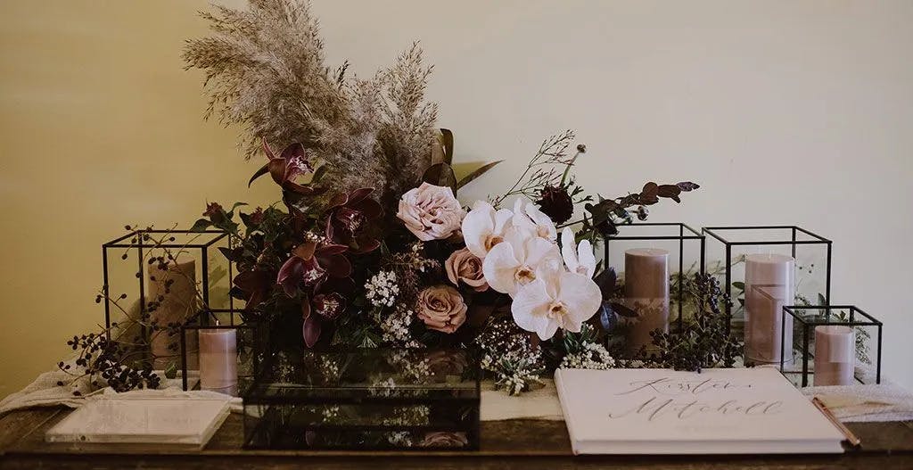 A wedding decoration arrangement featuring a variety of flowers, including white orchids, pink roses, and dark purple blooms, surrounded by greenery. Transparent boxes and candles are placed around the floral display, and a guestbook with the names "Krista and Mitchell" is open.