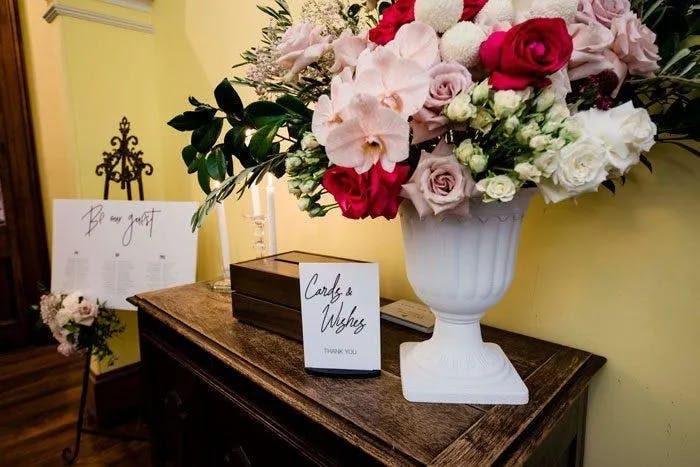 A wooden table adorned with a large white vase of colorful flowers, including red, white, and pink blossoms. Next to the vase is a sign labeled "Cards & Wishes, Thank You," and a black box. A stand in the background holds a sign with handwritten text.