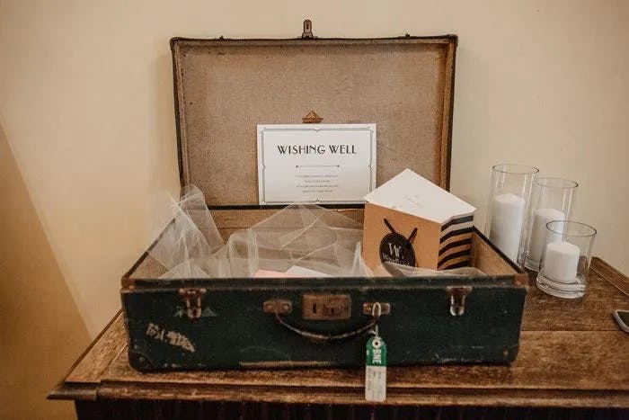 An old, open suitcase serves as a "wishing well" at an event, placed on a wooden table. It contains mesh bags and cards. Behind the suitcase are three tall glass candles, and a sign reading "Wishing Well" explaining its purpose.