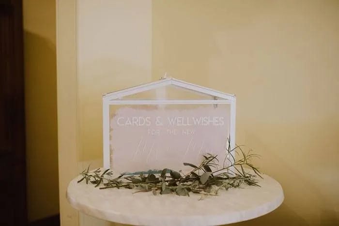 A white decorative box is placed on a small round table, with a sign reading "Cards & Well Wishes for the New Mr. & Mrs." Eucalyptus branches are arranged at the base of the box, all set against a pale yellow background.