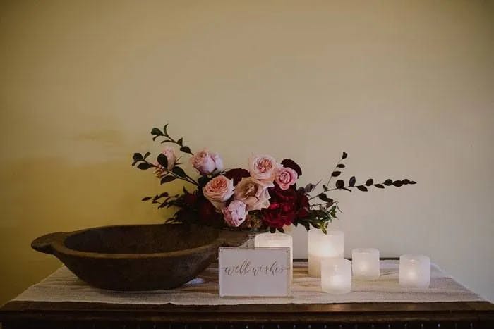A rustic wooden bowl with a floral arrangement of pink and red roses sits on a wooden table against a beige wall. To the right of the bowl, there are five lit candles of varying heights. In front of the bowl is a card that reads "well wishes.