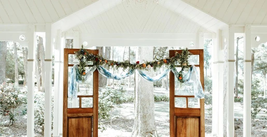 A rustic wedding arch setup with two wooden doors, adorned with green foliage, white flowers, and blue drapery, is placed under a white pavilion. The background shows a serene, wooded area with sunlight filtering through the trees.