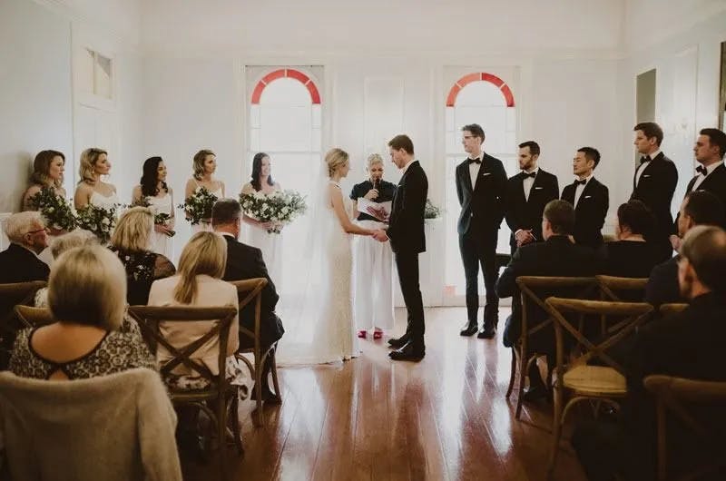 A bride and groom stand facing each other holding hands during a wedding ceremony with a bridal party beside them. The bride is in a white dress, and the groom is in a black suit. Guests are seated and watching attentively in a room with wooden floors and arched windows.