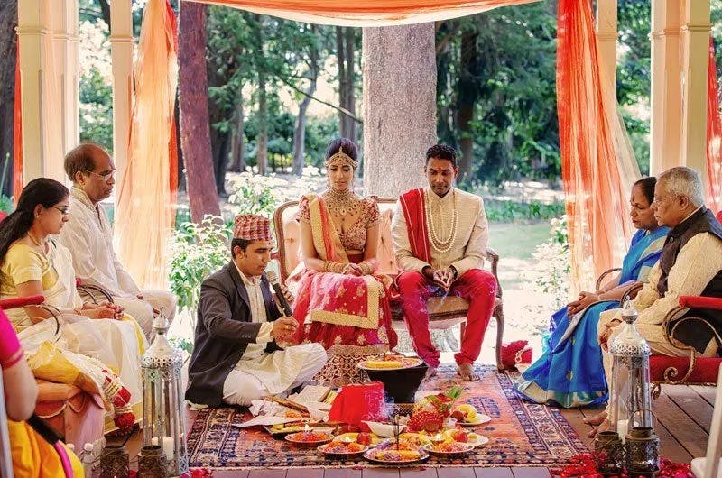A couple dressed in traditional attire sits under a decorative canopy during a wedding ceremony. They are surrounded by family members, with an officiant in a traditional outfit performing rituals. The scene features vibrant colors and traditional decorations.