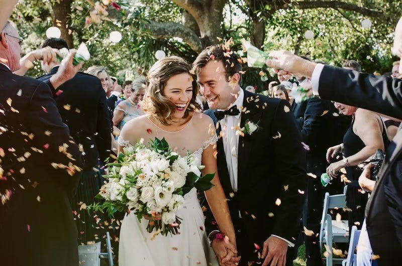 A newly married couple walks down the aisle, beaming with joy as guests throw confetti. The bride, in a white dress, holds a bouquet of white flowers and greenery. The groom, in a black suit and bowtie, smiles beside her. They are surrounded by happy friends and family.