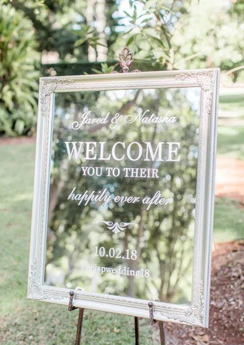 A glass wedding sign with white calligraphy sits on an easel in an outdoor setting. It reads: "Jared & Natasha WELCOME YOU TO THEIR happily ever after 10.02.18 #crispywedding18". The sign is framed with an ornate, white border. Lush greenery is in the background.