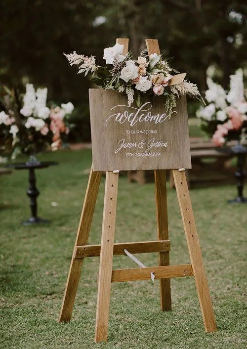 A wooden sign on an easel welcomes guests to a wedding, adorned with a floral arrangement of white and pink flowers. The sign reads, "Welcome to our wedding, James & Jessica, 19th October 2019." The background features greenery and more flowers.