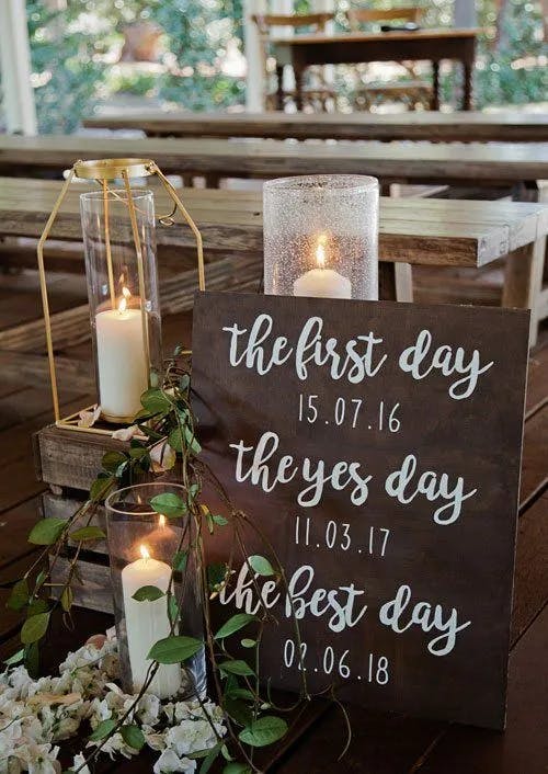 A wedding sign on a wooden floor with three dates: "the first day 15.07.16," "the yes day 11.03.17," and "the best day 02.06.18." The sign is surrounded by candles in glass holders, white flowers, and greenery. Wooden benches and foliage are in the background.