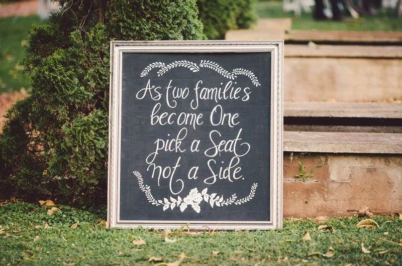 A chalkboard sign with a white ornate frame stands outdoors on grass next to a tree and brick steps. It reads, "As two families become one, pick a seat not a side," decorated with floral and leafy designs.