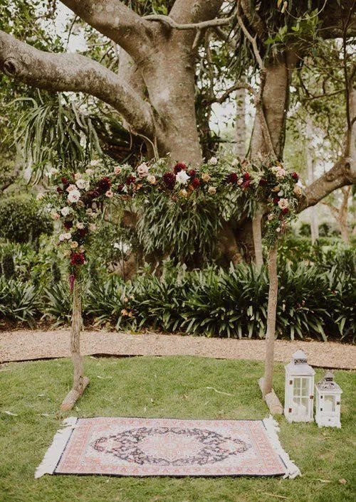 An outdoor wedding setup featuring a floral arch adorned with red and white flowers, set under a large tree. Below the arch is a patterned rug on grass, and two white lanterns are placed to the right of the rug, all surrounded by lush greenery.