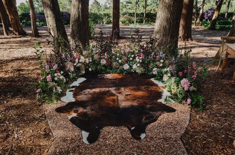 A wedding ceremony setup in a forest features a cowhide rug placed on the ground among tall trees. The rug is surrounded by a semicircle arrangement of colorful flowers and greenery. Wood chips cover the ground, adding to the rustic natural setting.