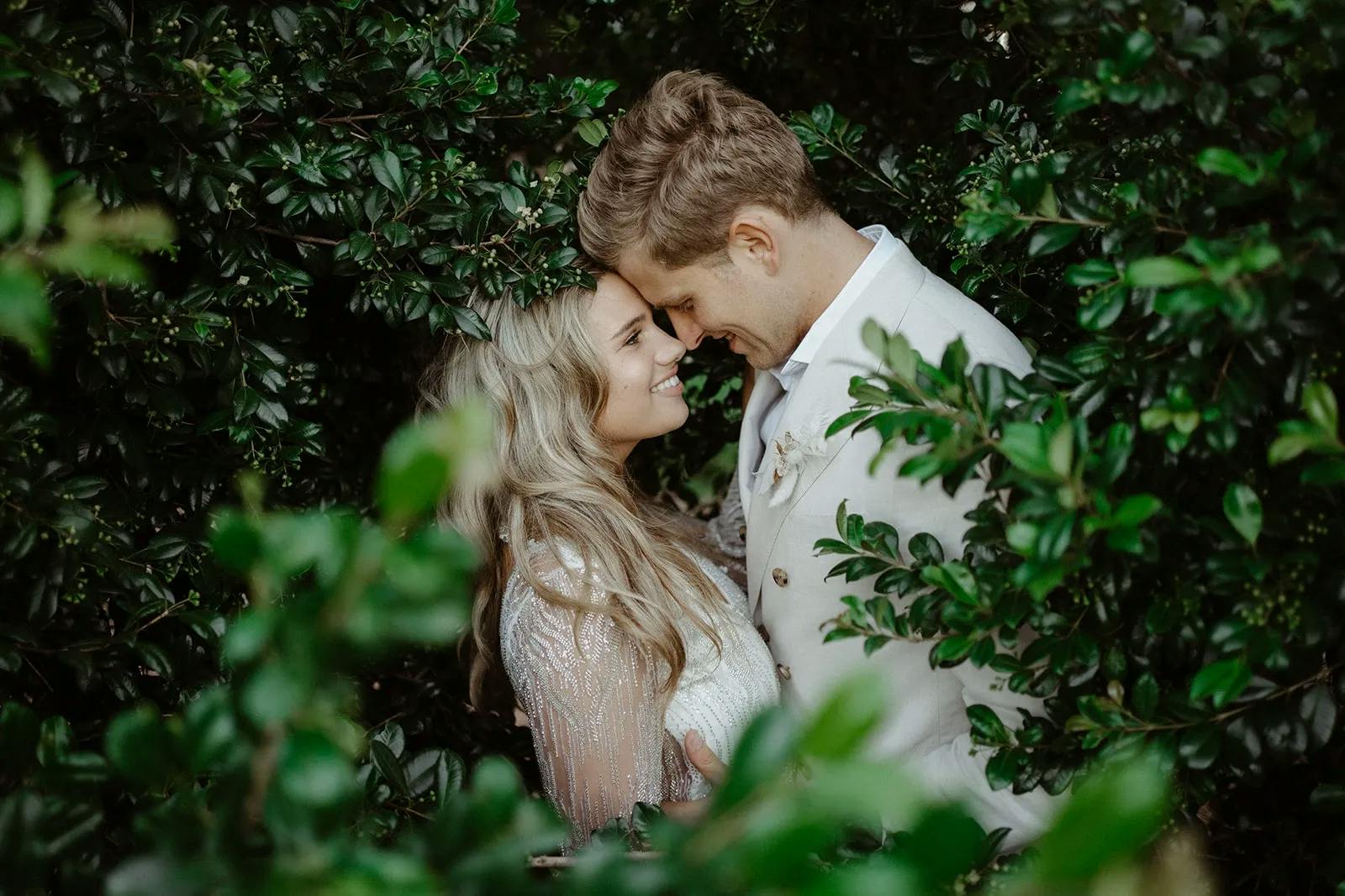 A couple shares an intimate moment, embraced among lush greenery. The woman, with long blonde hair, gazes lovingly at the man who is wearing a light-colored suit. The foliage surrounds them, creating a serene, romantic atmosphere.