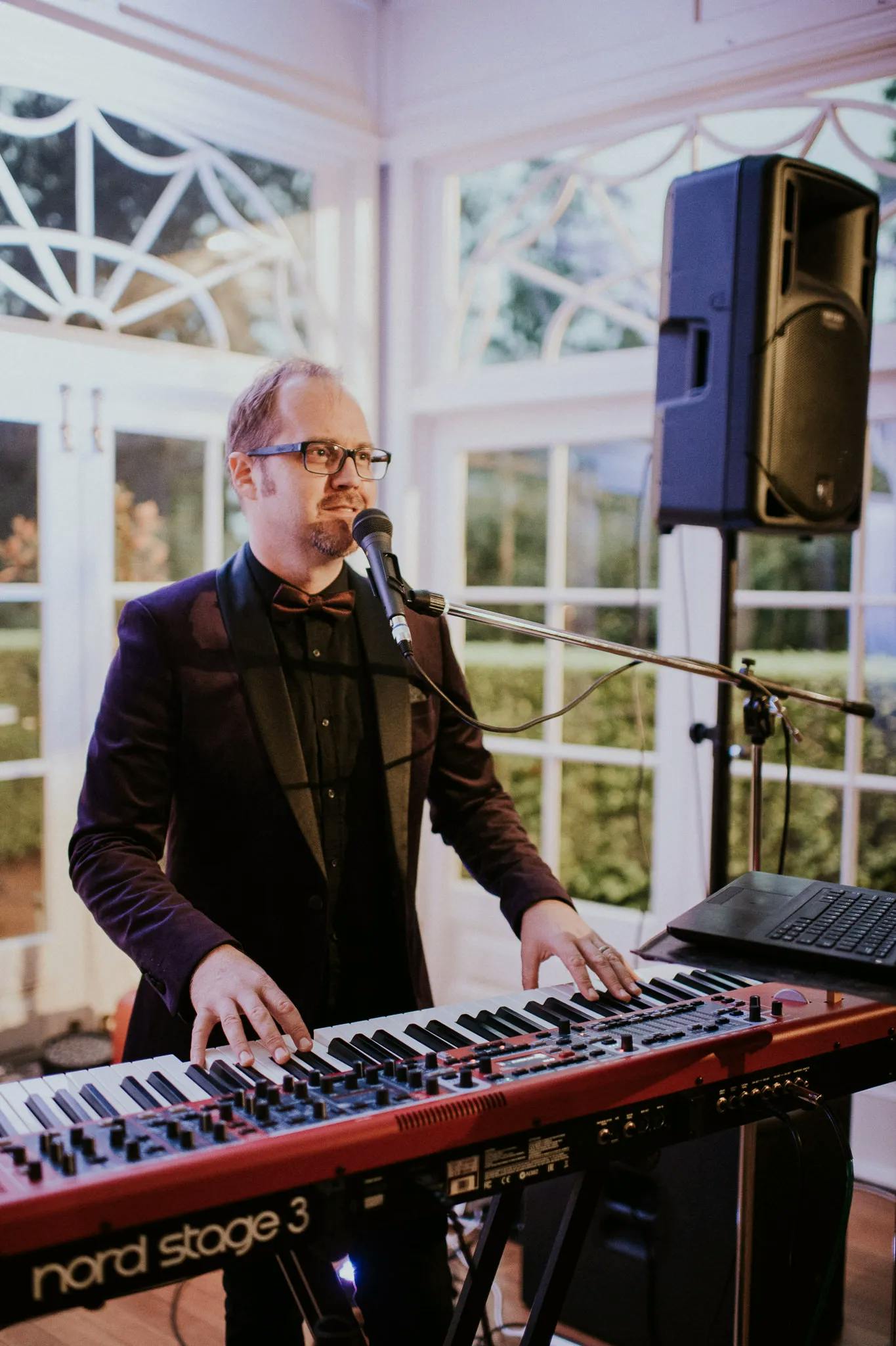A musician wearing glasses and a dark suit plays a Nord Stage 3 keyboard while singing into a microphone. He is performing in a well-lit room with large windows and a speaker positioned behind him.