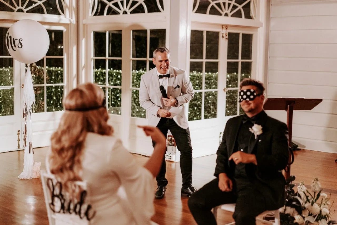 A bride and groom are playing a game indoors, with the groom blindfolded. The bride is smiling and pointing at him, while a man in a white jacket stands between them, holding a microphone and laughing. In the background, there are large windows and a "Mrs" balloon.