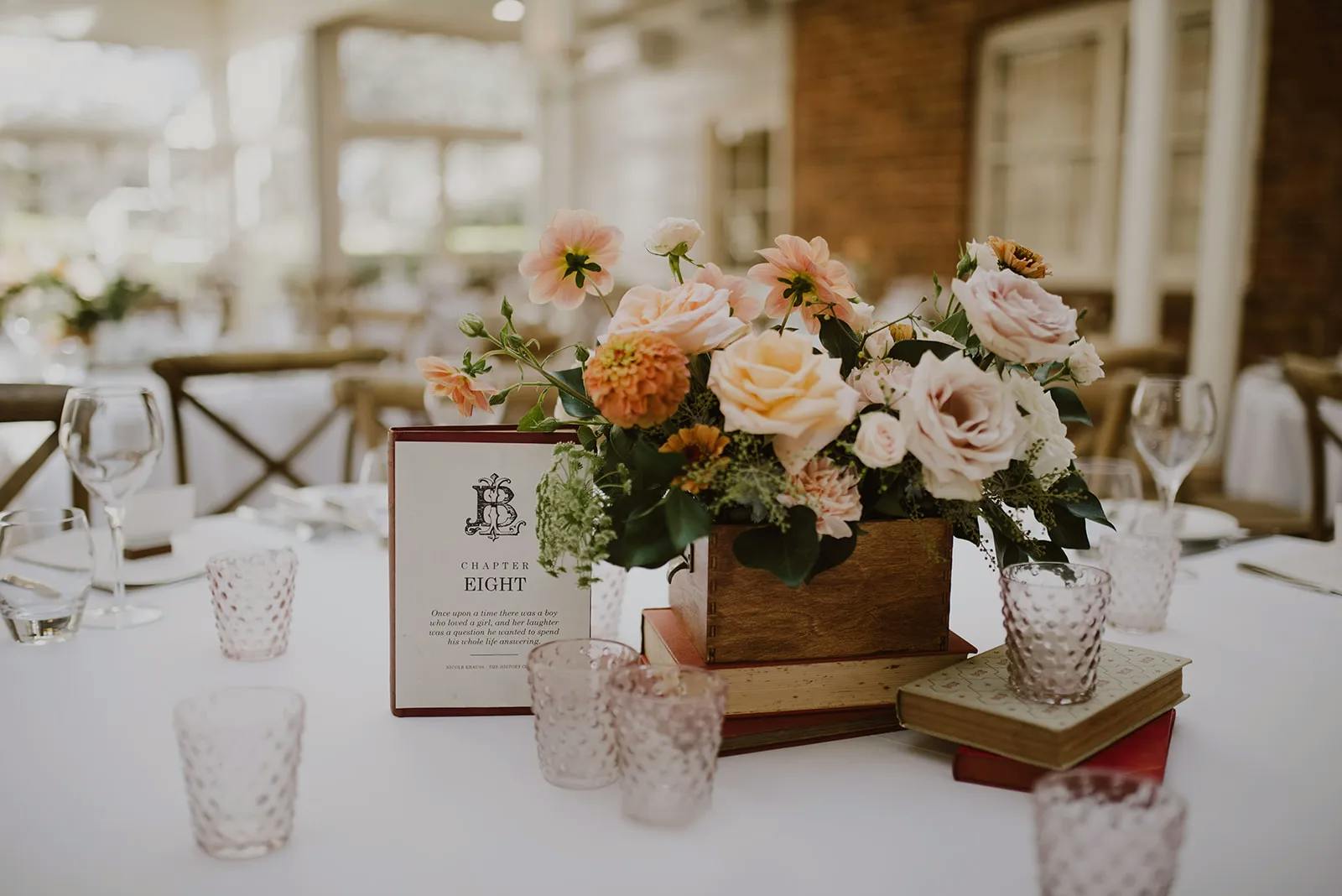 A table is elegantly set for an event, featuring a centerpiece of peach and orange flowers in a rustic box. Next to the arrangement is a standing card with "Chapter Eight" printed on it and some text. The table is adorned with pink glassware and vintage books.