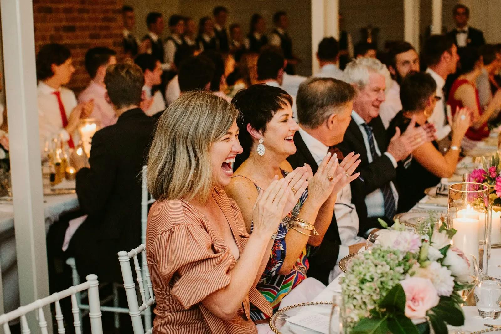 A group of people seated at a long, elegantly decorated table, smiling and clapping. The atmosphere is lively and festive, with floral centerpieces and candles. A brick wall and tuxedo-clad staff are visible in the background.