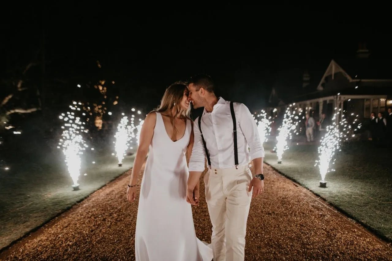 A couple, dressed elegantly with the woman in a white gown and the man in a white shirt and suspenders, walks hand in hand down a path lined with sparkling fireworks. They lean in for a kiss, surrounded by the romantic glow of the night and the vibrant fireworks.