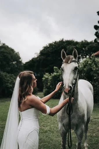 A bride in a white strapless wedding dress with a long veil gently touches the face of a white horse. They are outside in a lush green area with trees and bushes in the background, under a cloudy sky.