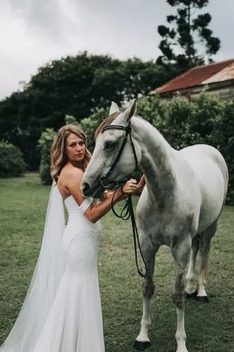 A woman in a white wedding dress stands next to a gray horse, holding its reins. They are outdoors on a lush, green lawn with trees and an old, rustic building in the background. The sky is overcast, creating a serene atmosphere.