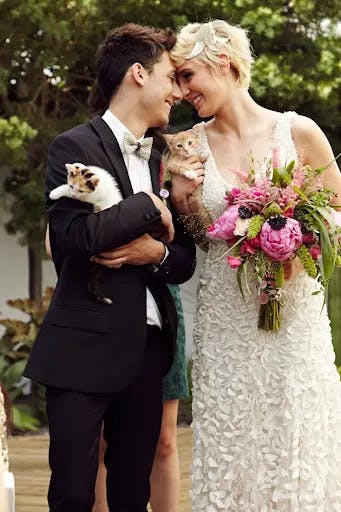 A newly married couple, dressed in a suit and a floral-patterned wedding dress, share a joyful moment while each holding a kitten. The bride also holds a vibrant bouquet of flowers, and they are standing outdoors, surrounded by greenery.