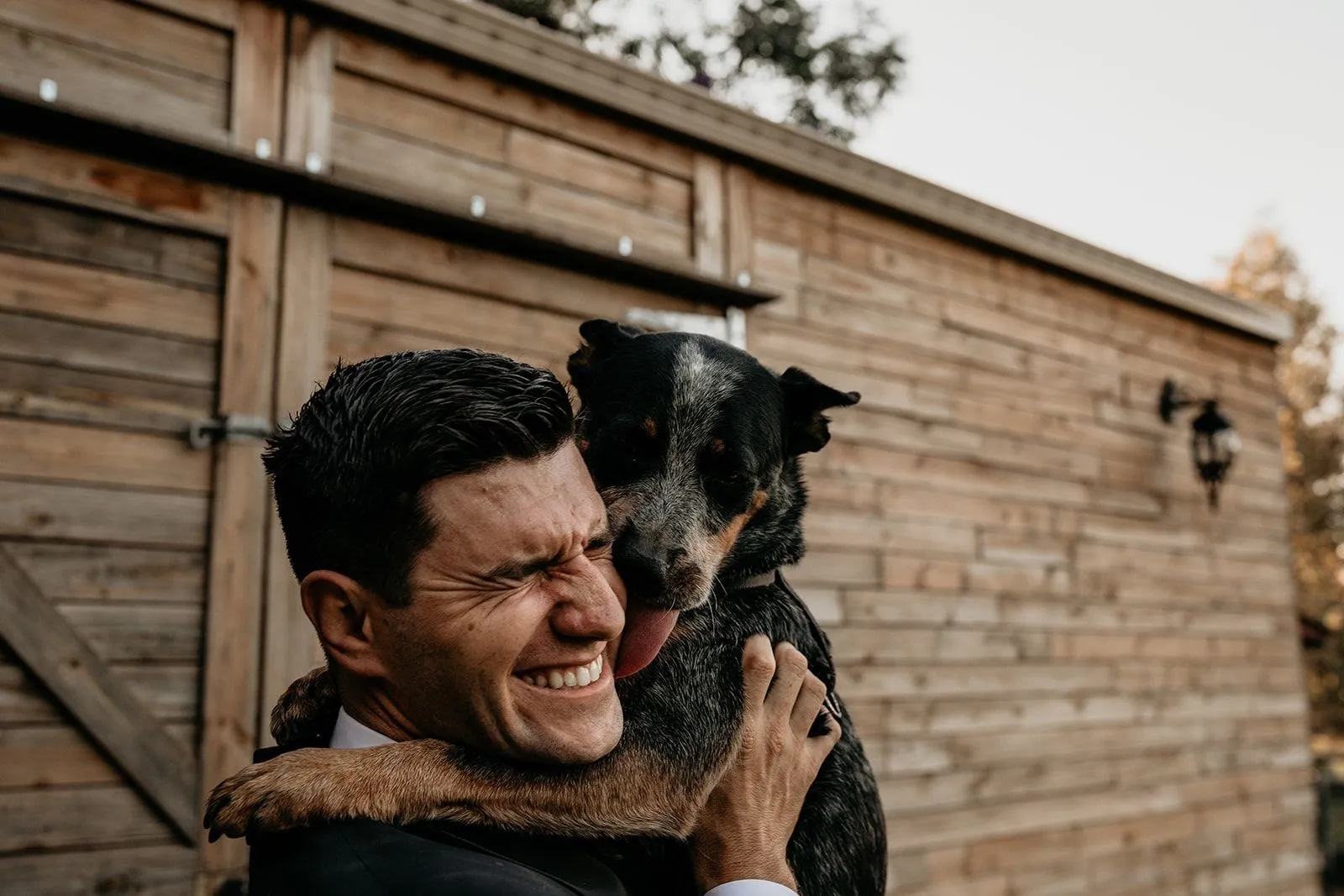 A man in a suit is hugging a dog that is licking his face, both looking joyous. They are standing outside in front of a wooden wall with a door. The man has short dark hair, and the dog has black and tan fur.