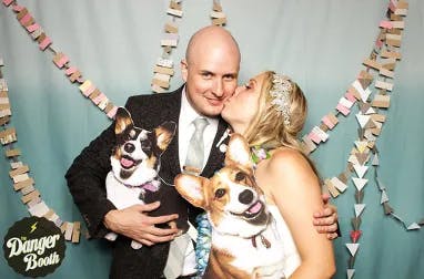 A man in a suit and a woman in a white dress pose against a decorated backdrop, holding dog-themed pillows. The woman is giving the man a kiss on the cheek. The backdrop features colorful paper garlands. A logo in the bottom left reads "Danger Booth.