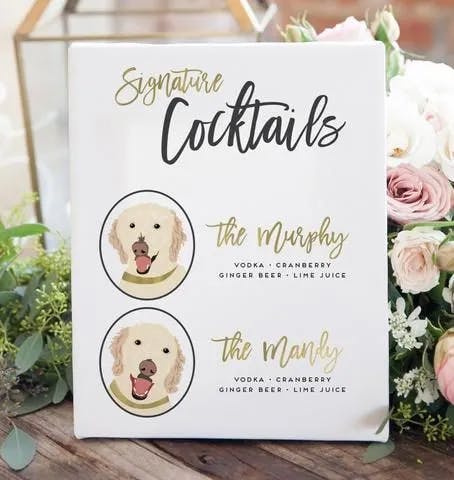 A wedding sign displays "Signature Cocktails" at the top with drawings of two dogs below. The drink names are "The Murphy" and "The Mandy," both containing vodka, cranberry, ginger beer, and lime juice. The sign is placed among floral arrangements.