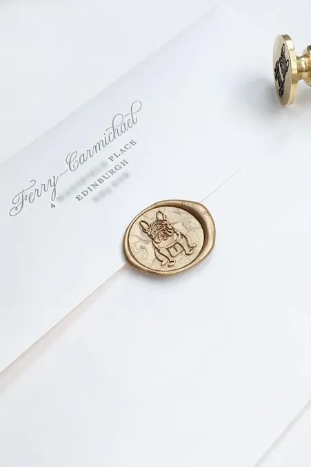 Close-up of a white envelope sealed with a gold wax stamp featuring an illustration of a figure. The envelope is addressed to "Terry Carmichael" with additional details indicating "4 PLACE EDINBURGH." A wax seal stamp tool is partially visible next to the envelope.