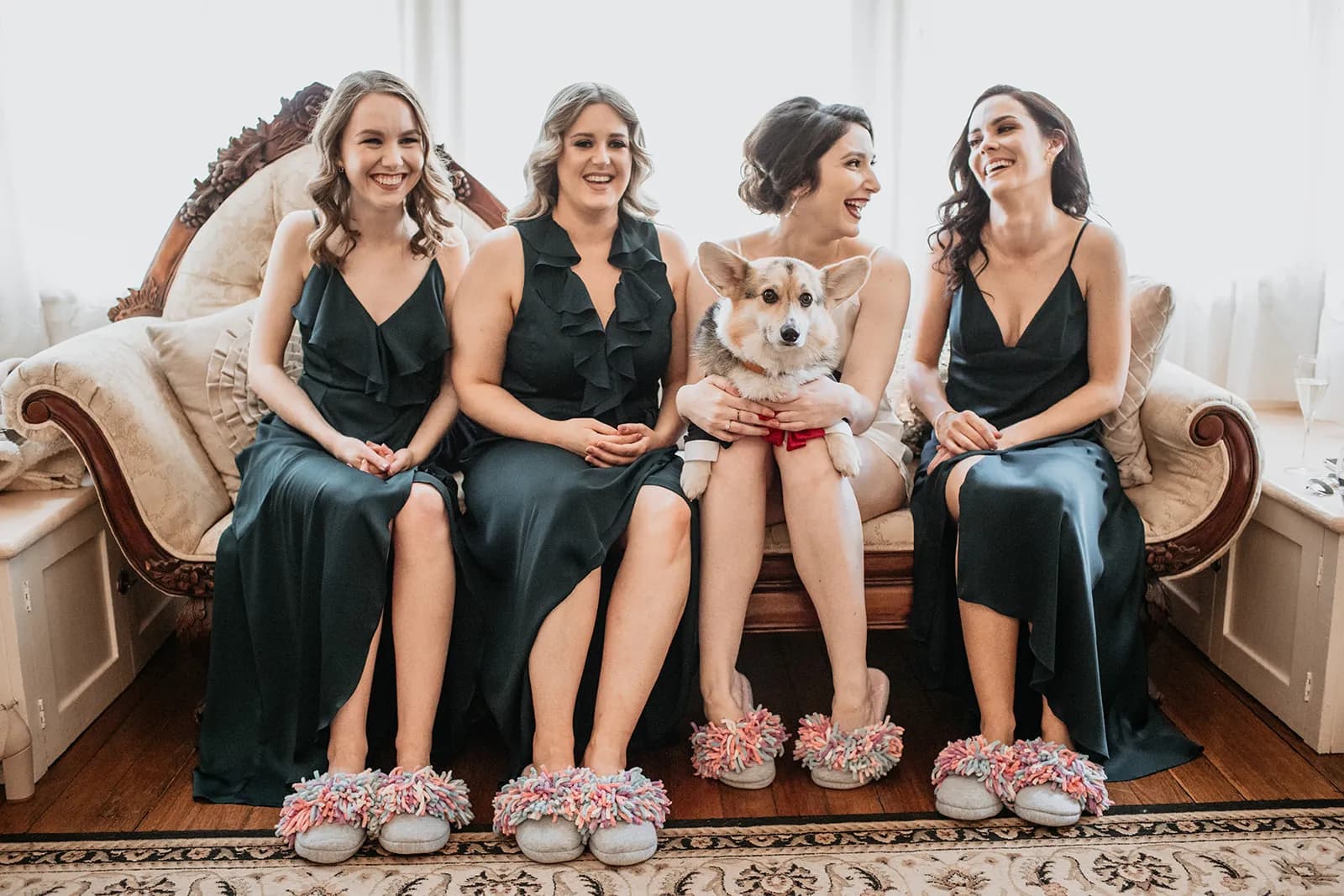 Four women in dark green dresses sit on an ornate couch. They wear matching multi-colored, fluffy slippers. One woman is holding a corgi wearing a red bow tie. They are all smiling, with a light background creating a warm atmosphere.