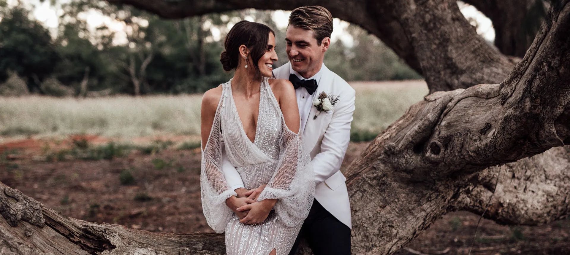 A couple, dressed in wedding attire, sits on a large tree branch. The bride wears a sparkling white gown with a deep V-neck and sheer sleeves, while the groom wears a white tuxedo with a black bow tie. They are looking at each other and smiling, surrounded by nature.