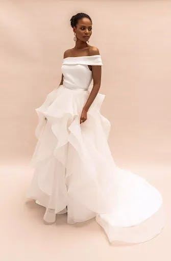 A woman stands against a plain background, wearing an elegant off-the-shoulder white wedding gown. The dress features a fitted bodice and a voluminous skirt with flowing, ruffled layers, and a long train. She has her hair styled in an updo and wears large, dangling earrings.