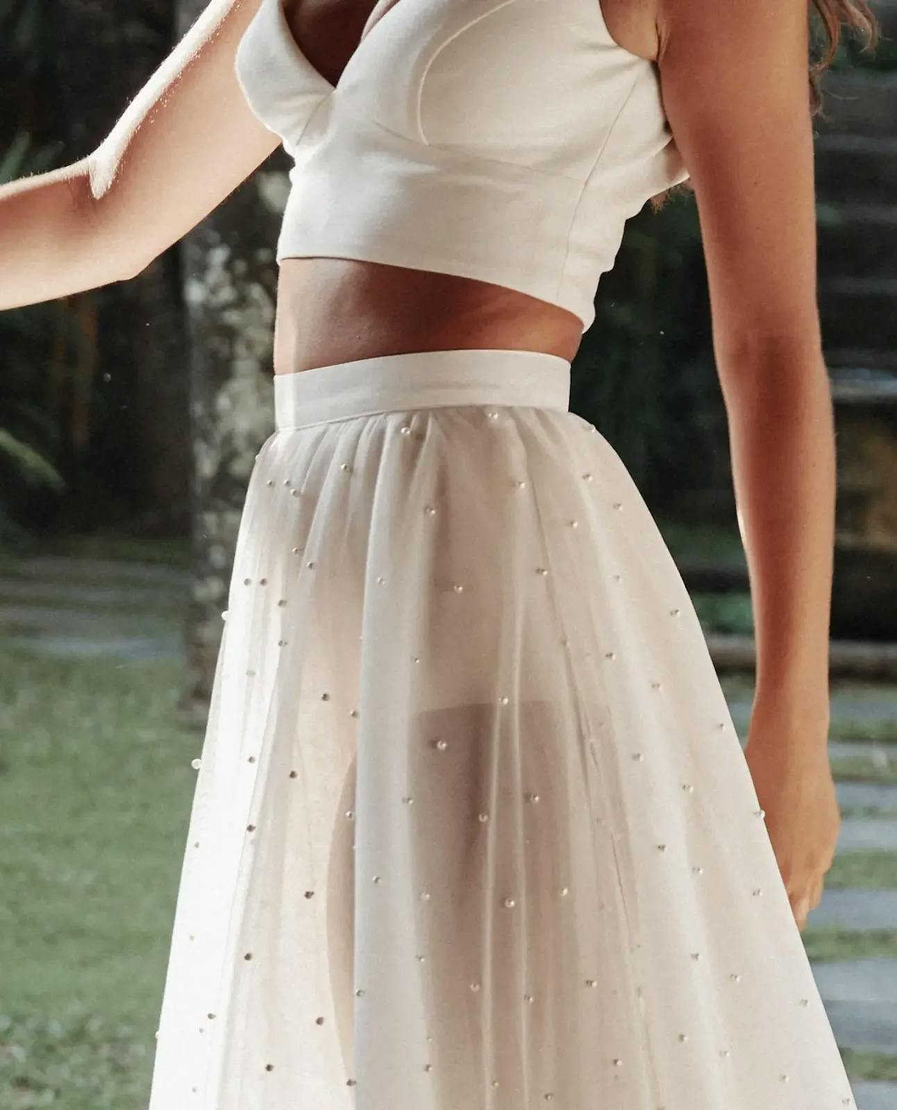 A woman is wearing a white two-piece outfit. The top is a sleeveless crop top with a deep neckline, and the bottom is a high-waisted, sheer, flowy skirt adorned with small, sporadic pearl-like beads. The background is an outdoor setting with green foliage.