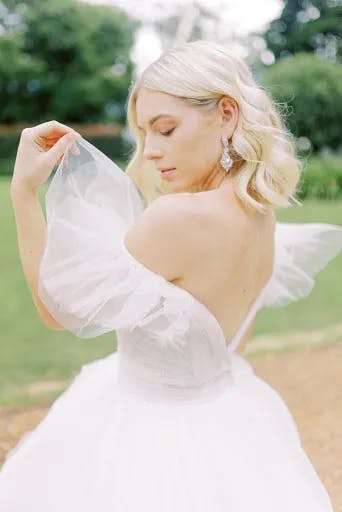 A woman with blonde hair wearing an elegant white off-shoulder wedding gown stands in an outdoor setting. She gently holds the sheer fabric of her dress, facing slightly away from the camera, with lush greenery in the background.
