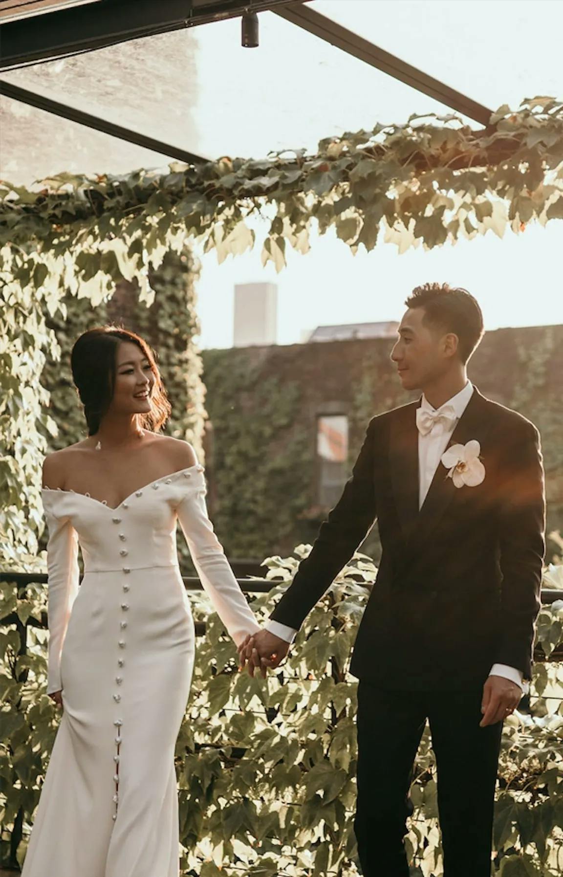 A bride and groom stand outdoors holding hands, both smiling at each other. The bride is wearing an elegant off-shoulder white dress with buttons down the front, while the groom is in a black tuxedo with a white shirt and bow tie. They are surrounded by greenery.