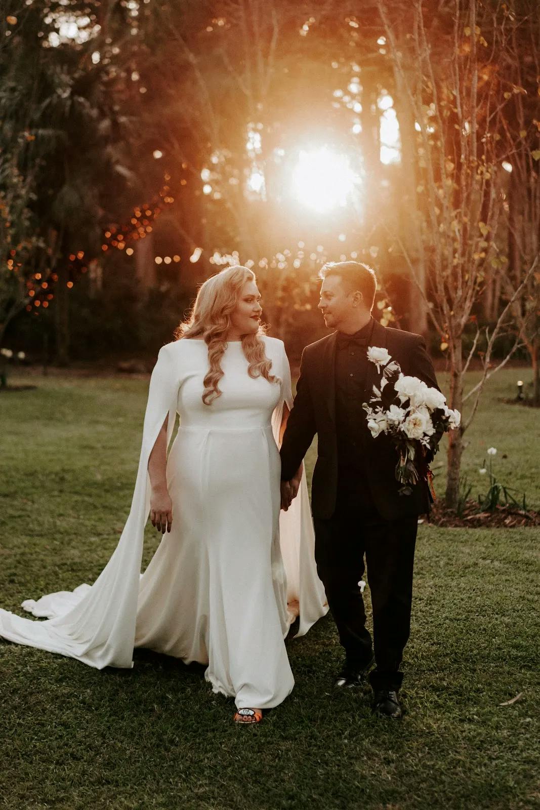 A woman in a white wedding gown with a long train and a man in a black suit holding a bouquet walk hand-in-hand on a grassy area. The setting sun filters through trees in the background, casting a warm glow and twinkling lights add a touch of magic to the scene.