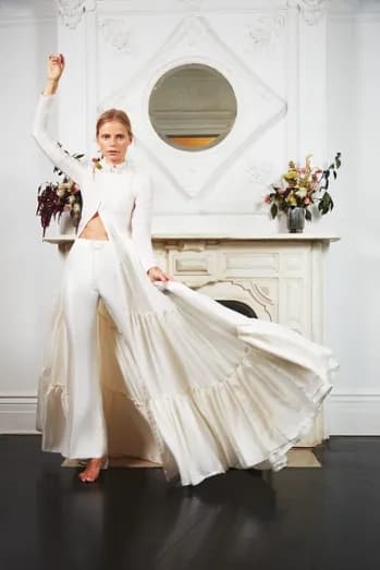 A woman in an elegant white outfit poses confidently indoors. She raises one arm while holding out the flowing skirt of her attire with the other. Behind her is an ornate white mantel with floral arrangements, and a round mirror hangs above it.