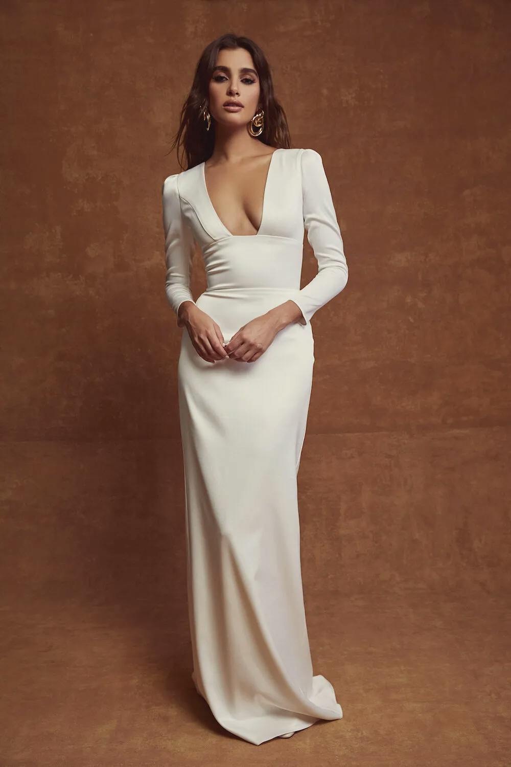 A woman stands confidently against a brown textured backdrop, wearing an elegant, long-sleeved, white evening gown with a deep V-neckline. Her dark hair is styled in loose waves, and she accessorizes with large, statement earrings.