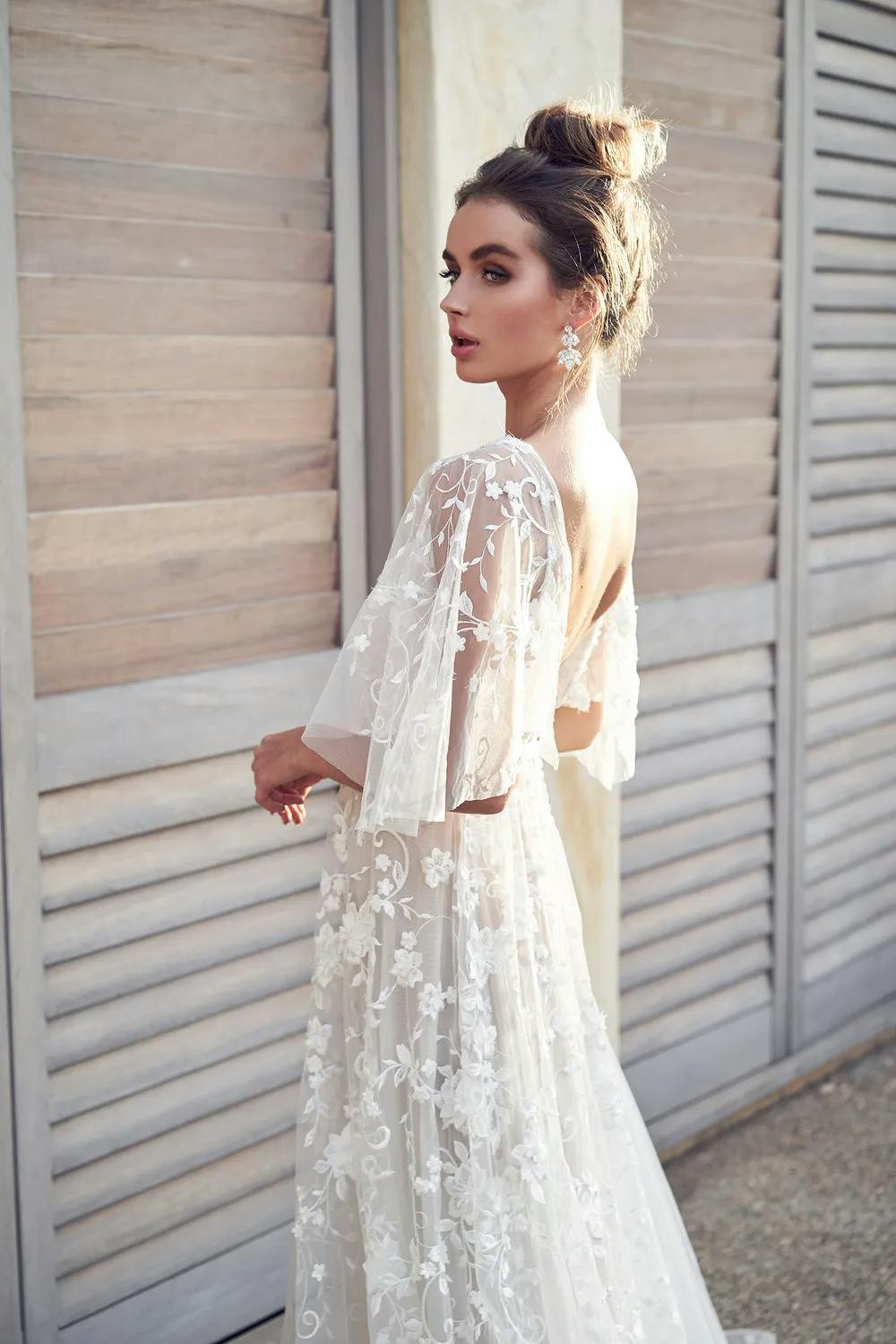 A woman stands in front of wooden shutters, wearing a white, sheer wedding gown with floral lace details. She has her hair in an updo and is turned slightly to the side, looking over her shoulder. She wears statement earrings, and the setting appears to be outdoors.