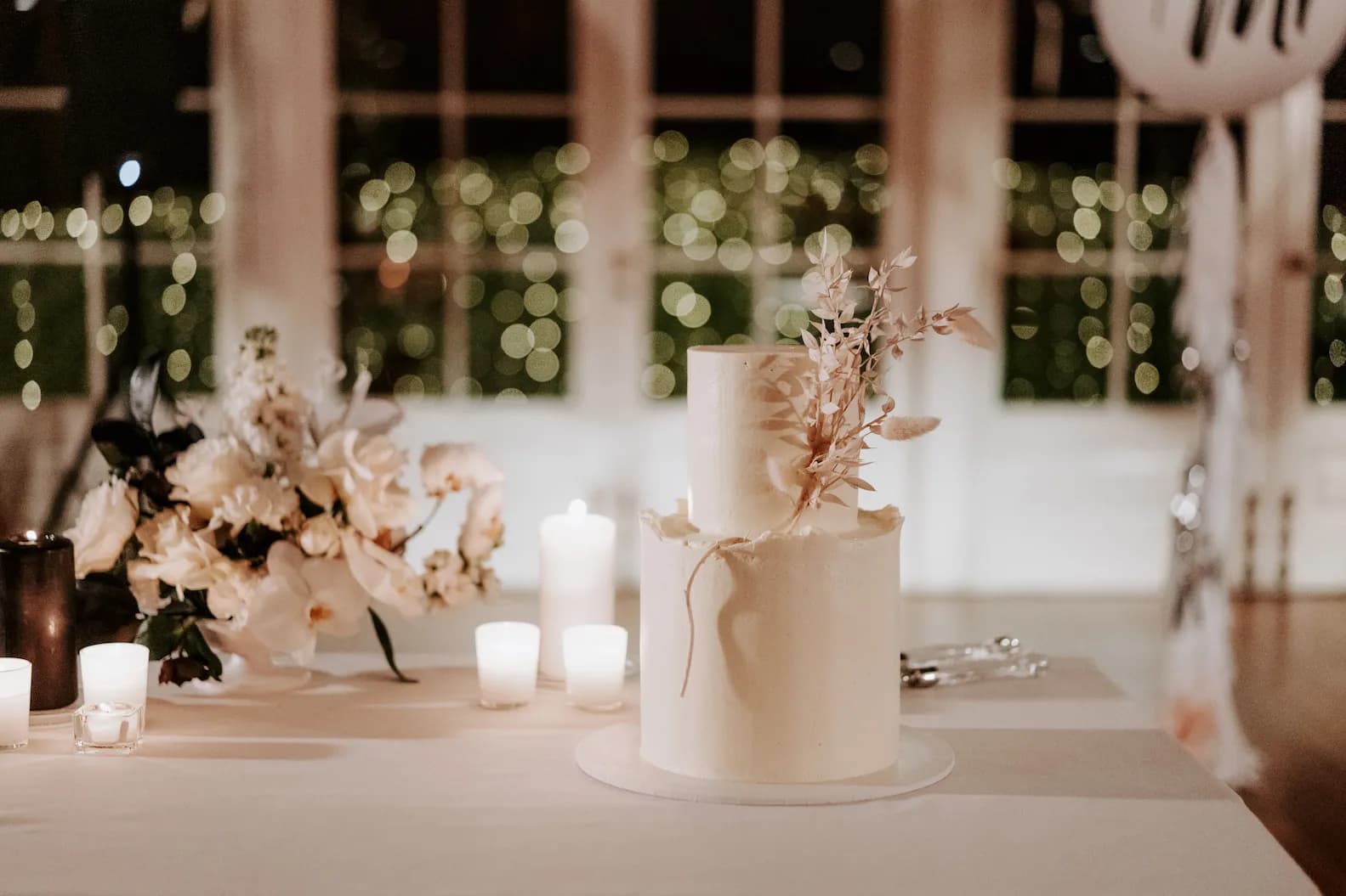 A white, two-tiered wedding cake with minimalistic floral decorations sits on a table adorned with lit candles and an elegant flower arrangement. Blurry lights twinkle in the background, creating a warm and romantic ambiance.