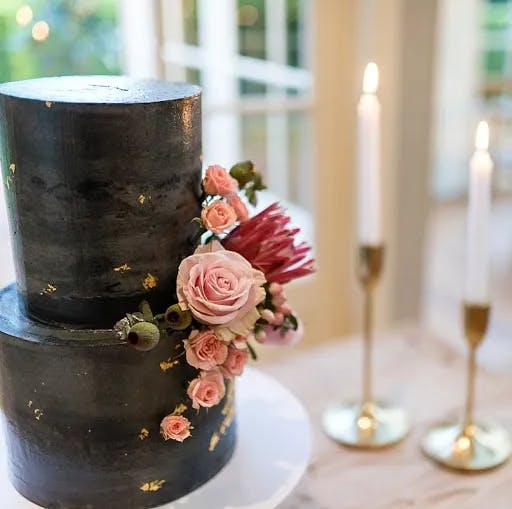 A tiered black wedding cake adorned with clusters of pink roses and other flowers is displayed indoors. The cake features flecks of gold foil. Two lit white taper candles in gold holders are placed in the blurred background on a marble surface.