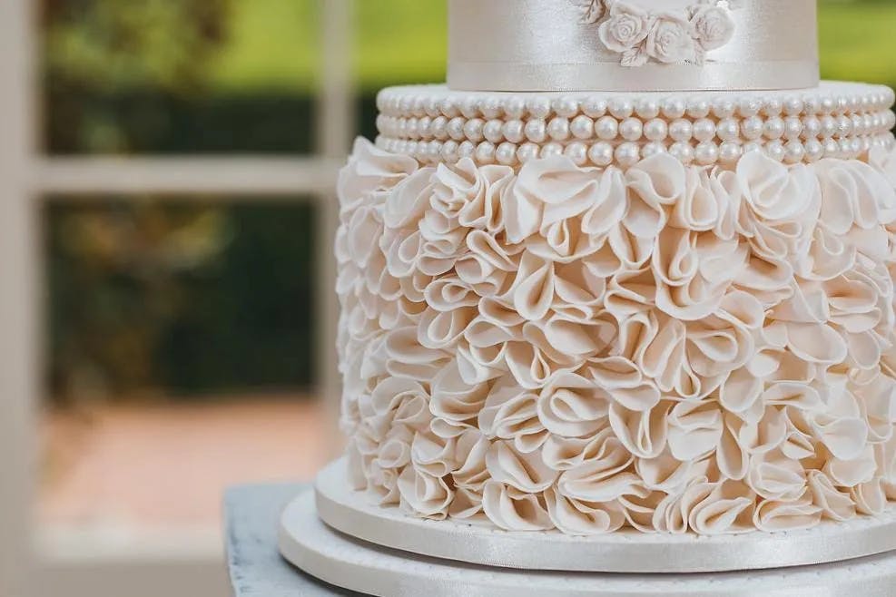 A close-up of an elegant white wedding cake. The cake is decorated with intricate ruffled icing that resembles delicate flower petals, adorned with a string of pearls, and white rose flowers on the top tier. The background shows a sunlit window with greenery outside.
