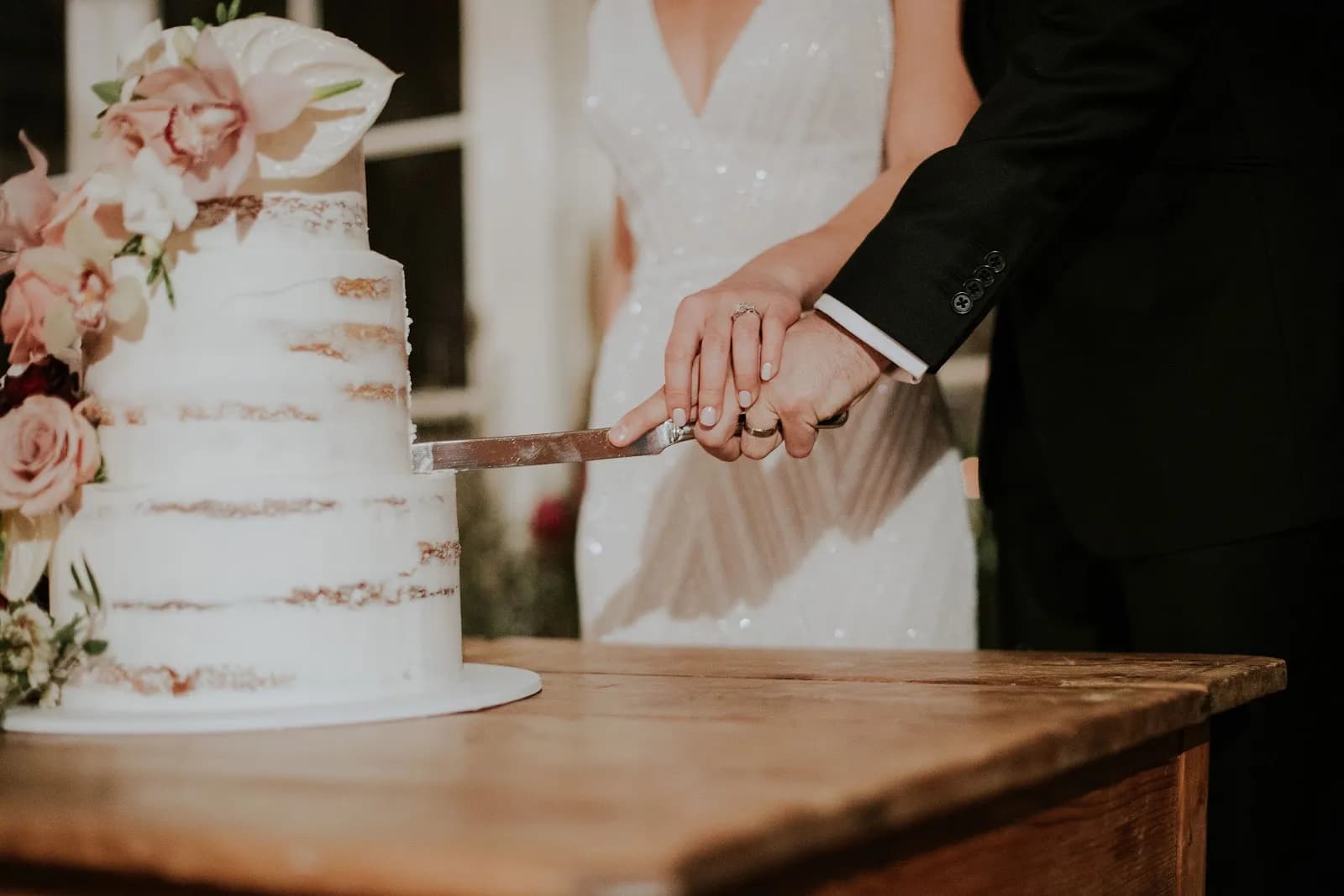 A bride and groom cut a wedding cake together with a knife. The cake is three tiers, semi-naked, decorated with flowers. The bride wears a white dress, and the groom wears a black suit. They are standing at a wooden table.