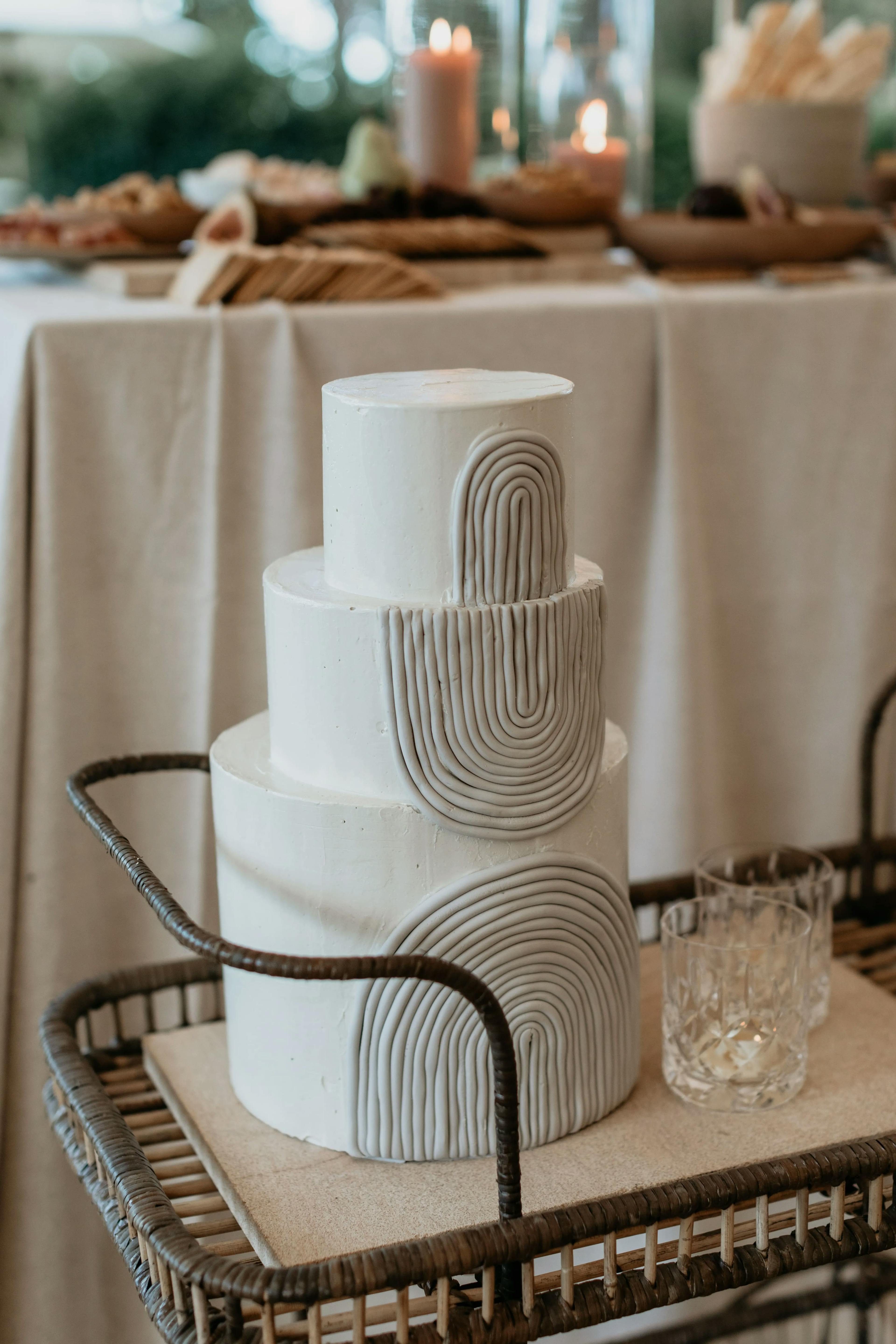 A three-tier white wedding cake with abstract textured icing designs sits on a wicker cart. Two empty glasses are placed next to the cake. In the blurred background, a table with candles and other food items is visible.
