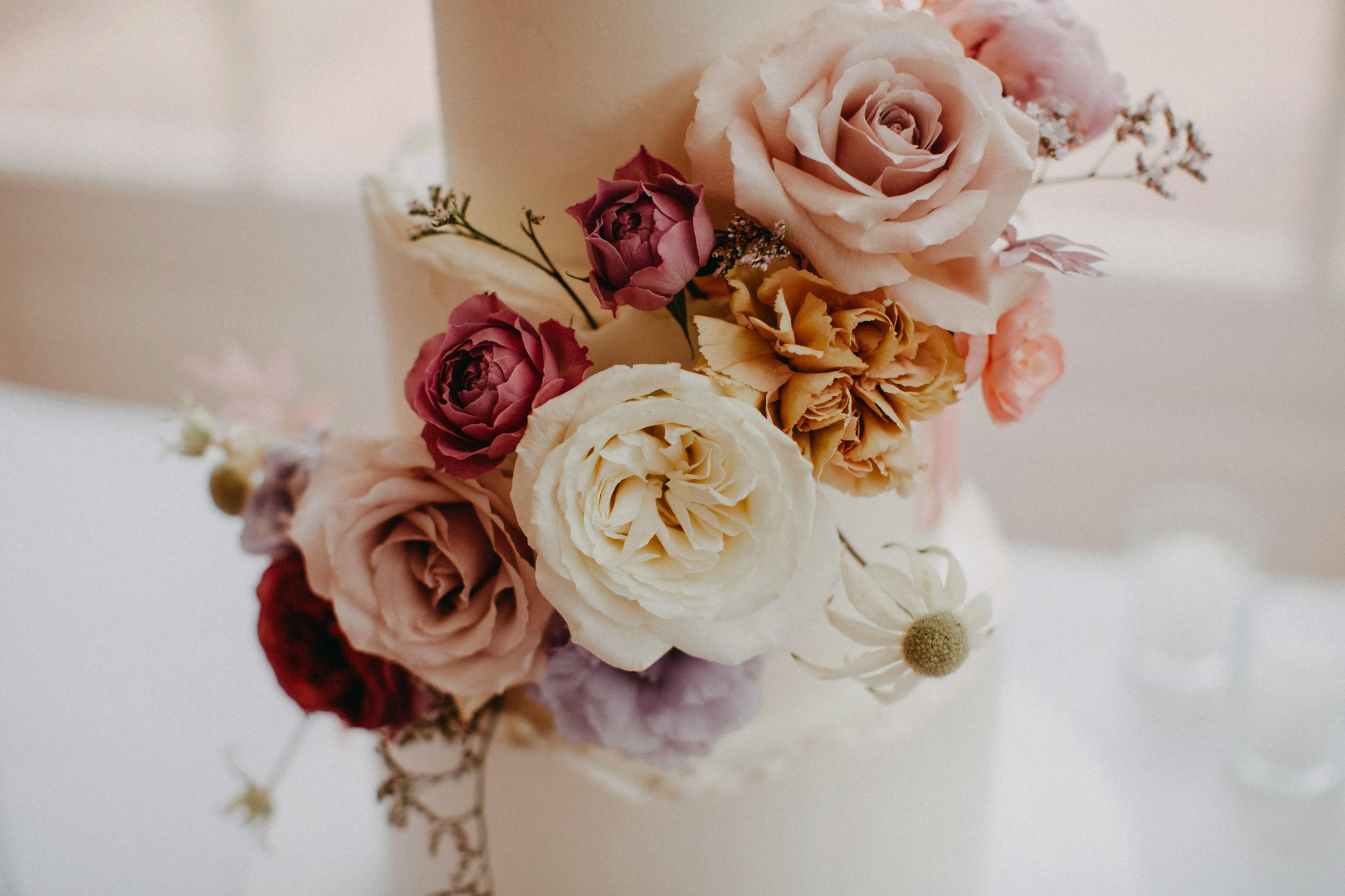 A two-tiered white wedding cake adorned with a beautiful arrangement of roses and other flowers in various colors including white, pink, and purple. The flowers cascade gently from the top to the bottom tier.