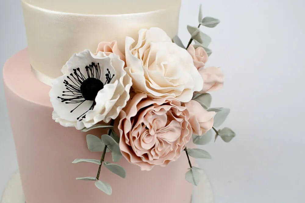 A two-tiered cake with a smooth, blush pink base and a subtle off-white top. The cake is beautifully adorned with elegant, realistic fondant flowers in soft shades of white and pink, complemented by delicate green leaves.