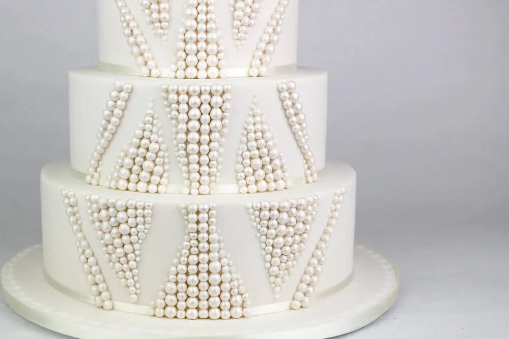 A three-tiered white wedding cake decorated with intricate geometric patterns made of pearl-like beads. The cake sits on a round white base against a plain gray background.