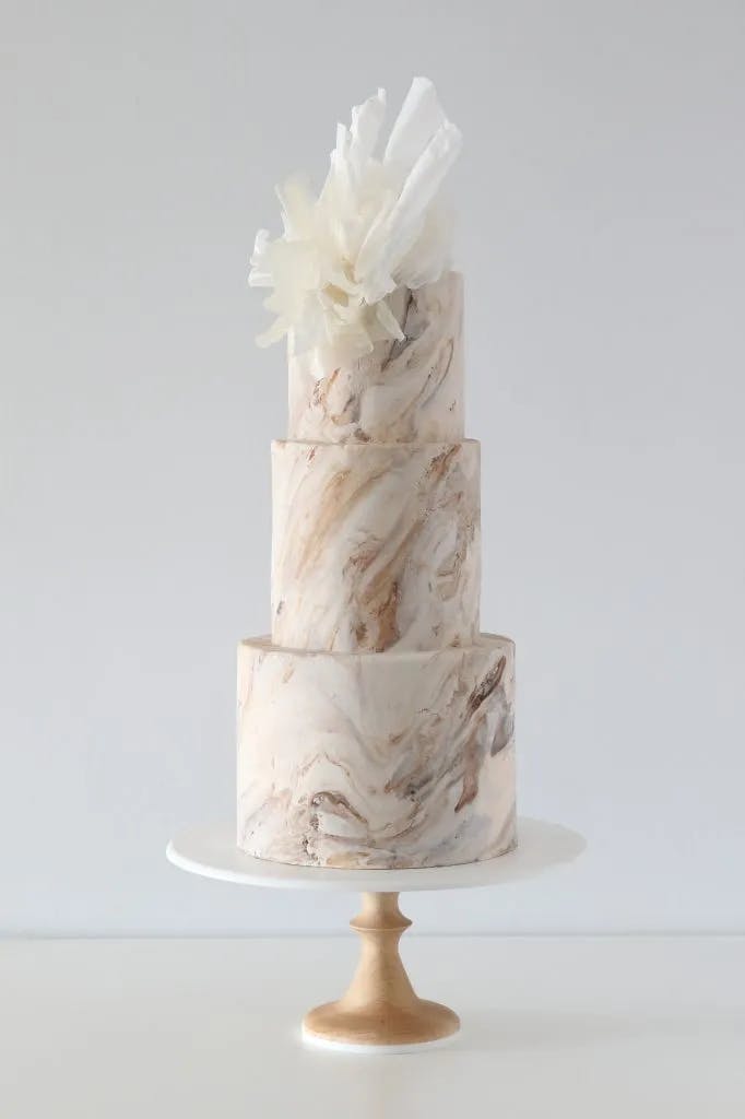 Three-tiered marble-themed cake on a wooden stand, featuring a white marbled design with light brown swirls. The top tier is adorned with large, delicate white sugar flowers, creating an elegant and modern appearance against a simple white background.
