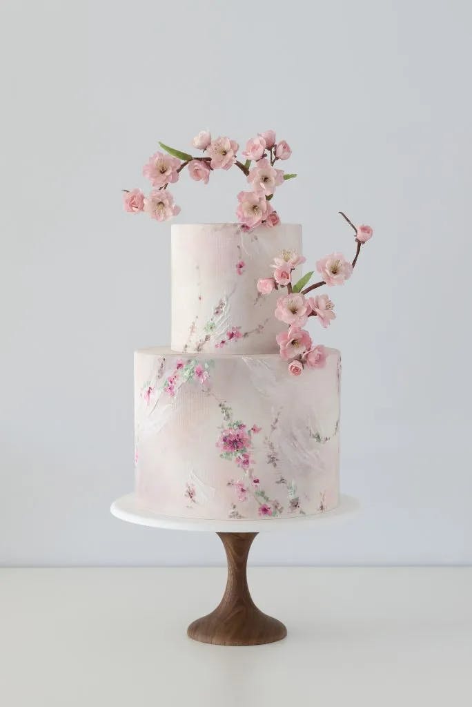 A two-tiered white cake adorned with delicate pink cherry blossoms. The cake features a rustic, textured finish and the blooms are arranged on top and cascading down the side. It is displayed on a sleek, wooden cake stand.